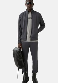 Ciemnoszara kurtka sportowa z wysokim kołnierzem, szary t-shirt, ciemnoszare spodnie, czarny plecak i czarne botki za kostkę. Gładki, aerodynamiczny design.