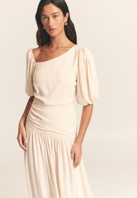 Bēšas krāsas kleita ar pūkainām īsām piedurknēm, vienas pleca dizains, savilkta vidukļa daļa un plūstoša svārku daļa. Gluda tekstūra, minimālisma stils.