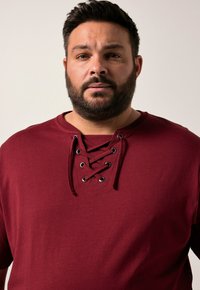 Bordeaux kortärmad skjorta med snörning framtill och svarta öglor, tillverkad av mjuktej s tyg med en slät yta.