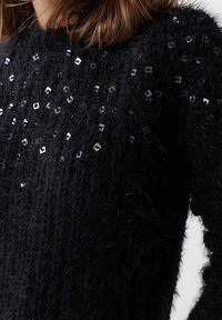 Černý chlupatý svetr s texturovaným pletením, na kterém jsou uspořádány čtvercové flitry ve směsici, což zdůrazňuje jeho prostorový vzhled.