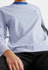 Vit långärmad skjorta med blå horisontella ränder, rund blå halsringning och diskret logotyp på bröstet, kombinerad med mörka byxor.