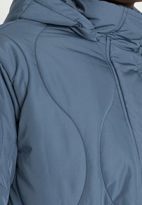 Chaqueta acolchada azul con capucha, que presenta una textura suave y un sutil detalle de costuras curvadas. Cierre de cremallera en la parte frontal visible.