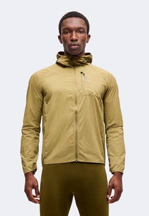 Mann trägt olivgrüne leichte Kapuzenjacke und dunkelgrüne Hose, steht frontal vor schlichtem hellem Hintergrund.