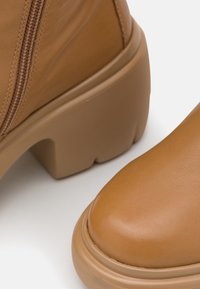 Bottes chevilles en cuir marron avec bout arrondi, talon épais et fermeture éclair latérale. Texture lisse et design minimaliste avec semelle plateforme.