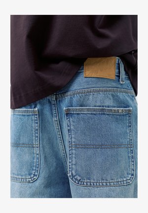 Vue arrière d'un jean en denim bleu clair avec deux poches arrière, une étiquette de marque en cuir marron et une chemise noire couvrant partiellement la ceinture.
