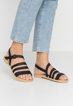 Black strappy sandals with a flat sole, featuring a buckle closure and a flexible rubber-like material. Worn with light blue cropped jeans.