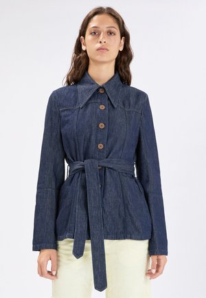 Donna che indossa una giacca di jeans blu scuro con colletto grande, bottoni in legno e cintura annodata in vita, abbinata a pantaloni chiari.