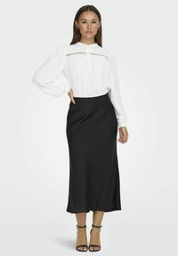 Blouse blanche à manches longues avec un col à bouton et un détail texturé, associée à une jupe midi noire en tissu lisse. Talons strappy noirs.