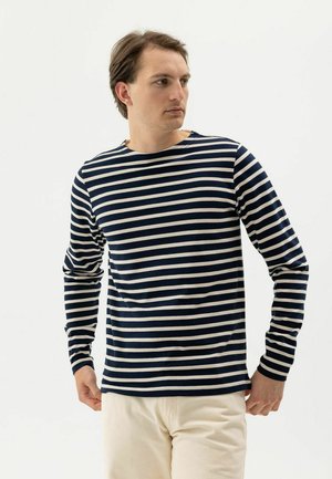 Férfi, aki tengerészkék-fehér vízszintes csíkos hosszú ujjú inget és világos bézs nadrágot visel, balra néz, egy egyszerű háttér előtt.