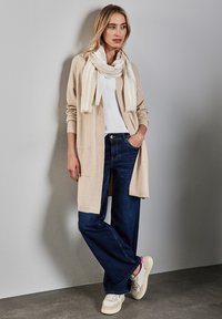 Beige cardigan, hvid tørklæde, hvid t-shirt, mørkeblå vide ben-jeans og off-white sneakers med brune accenter. Glatte teksturer og afslappet pasform.