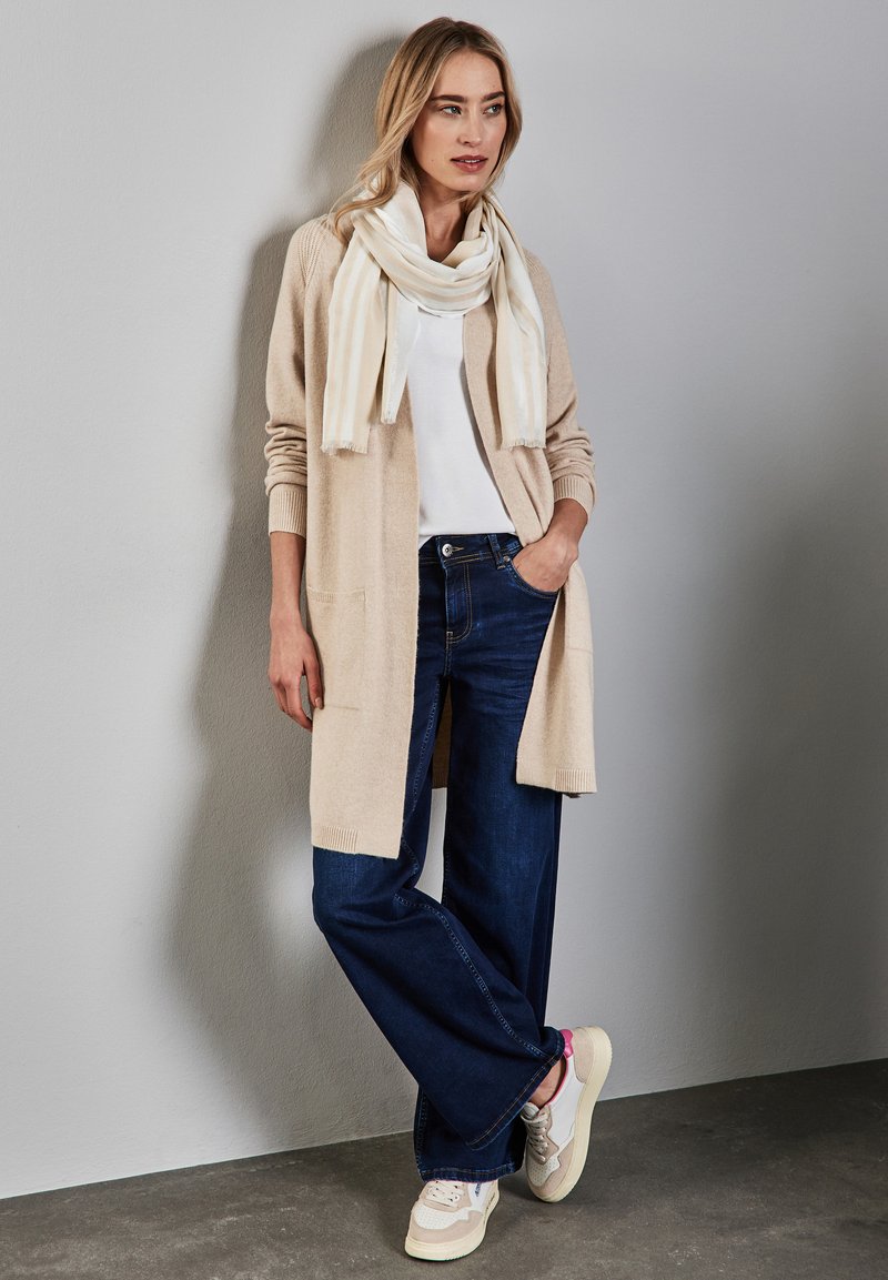 Beige cardigan, hvid tørklæde, hvid t-shirt, mørkeblå vide ben-jeans og off-white sneakers med brune accenter. Glatte teksturer og afslappet pasform.