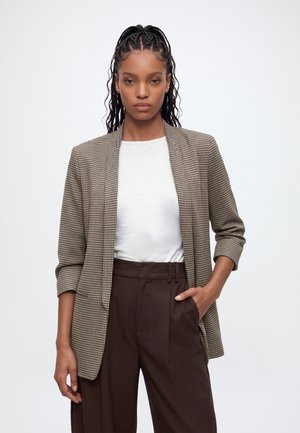 Femme aux cheveux tressés porte un blazer marron à carreaux, une chemise blanche et un pantalon taille haute marron foncé, posant avec une main dans la poche.