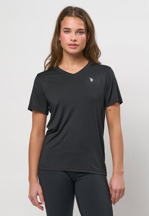 Mladá žena s dlhými vlasmi, oblečená v čiernom tričku s výstrihom do V a krátkym rukávom, s malým vyšívaným logom na hrudi, stojaca pred jednofarebným pozadím.