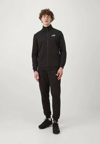 Młody mężczyzna stoi, ubrany w czarną bluzę z zamkiem marki Puma, dopasowane czarne dresy Puma oraz czarne trampki Puma, na jednolitym jasnym tle.