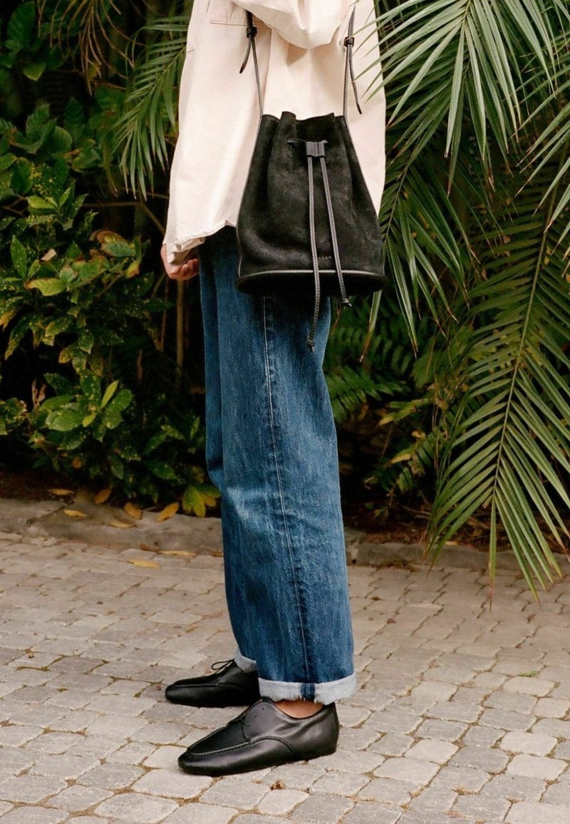 Personne portant un jean bleu retroussé, des chaussures en cuir noir et une veste légère, tenant un sac seau en cuir noir, debout sur un chemin pavé près de plantes vertes.