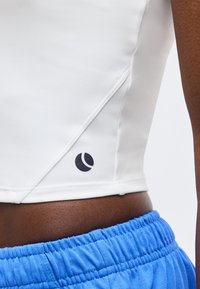 Κοντινό πλάνο της μέσης ενός ατόμου που φοράει λευκή εφαρμοστή μπλούζα με μικρό μαύρο λογότυπο και μπλε παντελόνι με ελαστική μέση.