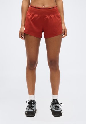 Αθλητής που φοράει κόκκινα αθλητικά σορτς, λευκές κάλτσες και μαύρα και γκρι παπούτσια τρεξίματος, στέκεται μπροστά σε απλό φόντο.