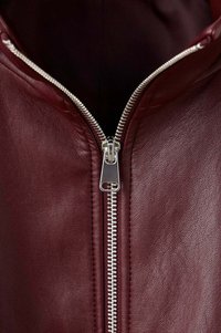 Fermeture éclair argentée partiellement fermée sur une veste en cuir bordeaux, montrant la surface texturée du cuir et la doublure intérieure foncée.