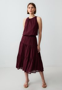 Abito a strati bordeaux con scollatura all'americana, caratterizzato da un corpetto arricciato e dettagli con bottoni lungo il lato, abbinato a sandali beige.