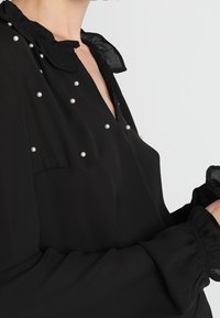 Mustaharja paita, jossa on teksturoitu, läpinäkyvä kangas. Rypytetty kaulus, jossa on helmikoristeita ja joustavat hihansuut. Irtonainen, tyylikäs muotoilu.