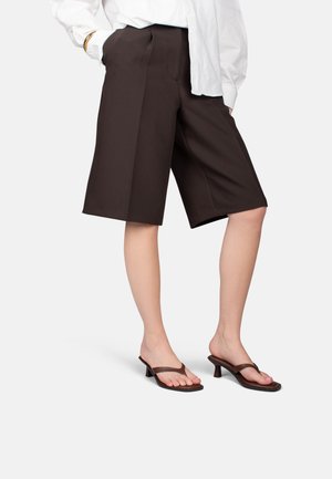 Femme portant un short ample marron foncé au genou, un chemisier blanc et des sandales à talons marron foncé avec brides, debout devant un fond uni.