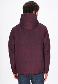 Burgundyröd puffjacka med huva, med ett texturerat mönster och elastiska muddar, som bärs över blå jeans, sedd bakifrån.