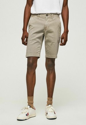 Pantaloni beige con una superficie texturizzata, chiusura con bottone e tasche laterali. Abbinati a sneakers bianche e calze color sabbia, in piedi su uno sfondo neutro.