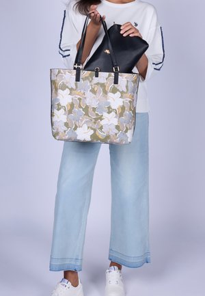 Person hält eine Tasche mit floralem Muster mit schwarzen Griffen und eine schwarze Clutch, trägt hellblaue Jeans und weiße Sneaker.