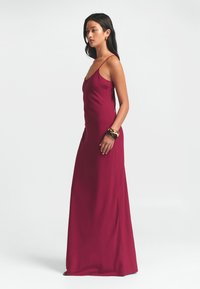 Burgundijos spalvos, grindis siekianti suknelė su plonomis petnešėlėmis ir lygia, prigludusia siluetu, su šoniniu įplėšimu ir minimalistiniais dizaino elementais.