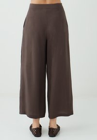 Brede broek in donkerbruin stof, met een hoge taille, zijzakken en een gladde textuur. De zoom valt boven de enkel.