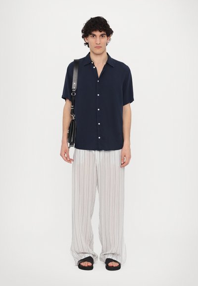 Chemise bleu marine à manches courtes, pantalon lâche à rayures blanches et sandales noires. La chemise présente un devant à boutons ; les pantalons ont une texture légère.