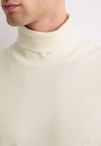 Cremfarbener Rollkragenpullover aus geripptem Stoff, mit hohem Kragen und glatter Textur, betont die figurbetonte Passform und die neutrale Farbgebung.