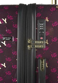 Maleta rígida DKNY en morado oscuro con un patrón geométrico en rosa y dorado. Presenta cremalleras negras, un candado de combinación y herrajes de marca.