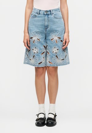 Riflové šortky po kolená s veľkými bielymi vyšitými kvetinovými výrezmi, nosené s bielymi ponožkami a čiernymi topánkami na pracku na stojacej osobe.