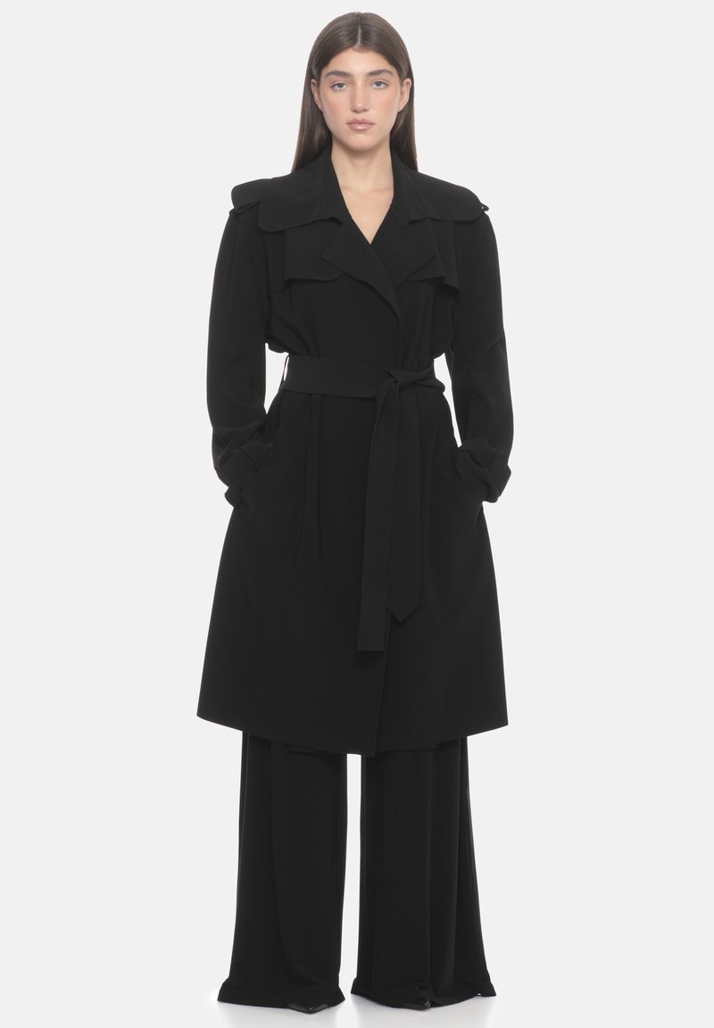 Trench-coat noir avec ceinture, larges revers, manches longues et poches latérales, présentant une texture de tissu lisse et une coupe ajustée.