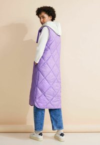 Lång lila vadderad väst med huva, diamantmönster, sidofickor, sliten över en ljusgrå tröja, kombinerad med blåjeans och sneakers.