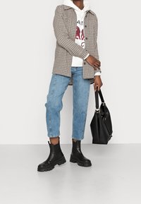 Camisa a cuadros marrón y beige, sudadera blanca con gráficos rojos, vaqueros azul claro, botas negras de tobillo con suelas gruesas y una bolsa tote negra.