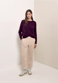 Frau, die mit einem dunkelvioletten Pullover, beigen Hosen mit Taschen und weißen Turnschuhen vor einem einfachen zweifarbigen beigen Wandhintergrund steht.