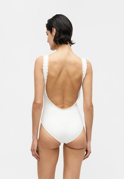 Γυναίκα με κοντά σκούρα μαλλιά που φοράει λευκό ολόσωμο μαγιό με βαθύ άνοιγμα στην πλάτη, στέκεται μπροστά σε ένα απλό φωτεινό φόντο.