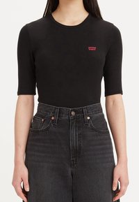 Žena v těsném černém tričku s krátkým rukávem a malým červeným logem Levi's a v tmavě šedých džínách s vysokým pasem na bílém pozadí.