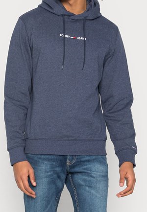 Homme portant un sweat à capuche Tommy Jeans bleu marine avec cordons et un jean bleu, debout les mains détendues le long du corps.