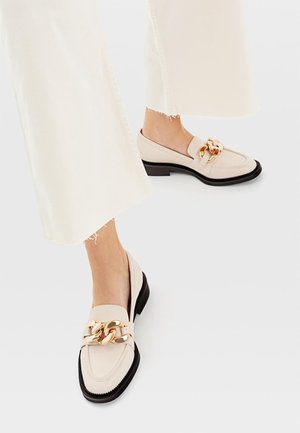 Beige læder loafers med en guldkæde accent, featuring en rund tå og lav sort hæl, parret med cremefarvede vidde bukser.