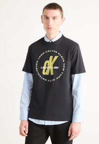 Melns kokvilnas T-krekls ar apļu grafiku, kurā uzraksts "Calvin Klein" ir baltā un zelta krāsā, apakšā valkāts ar gaiši zilu krekla jaku.