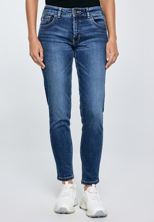 Žena u tijesnim plavim trapericama i bijelim tenisicama stoji uz običnu svijetlu pozadinu.