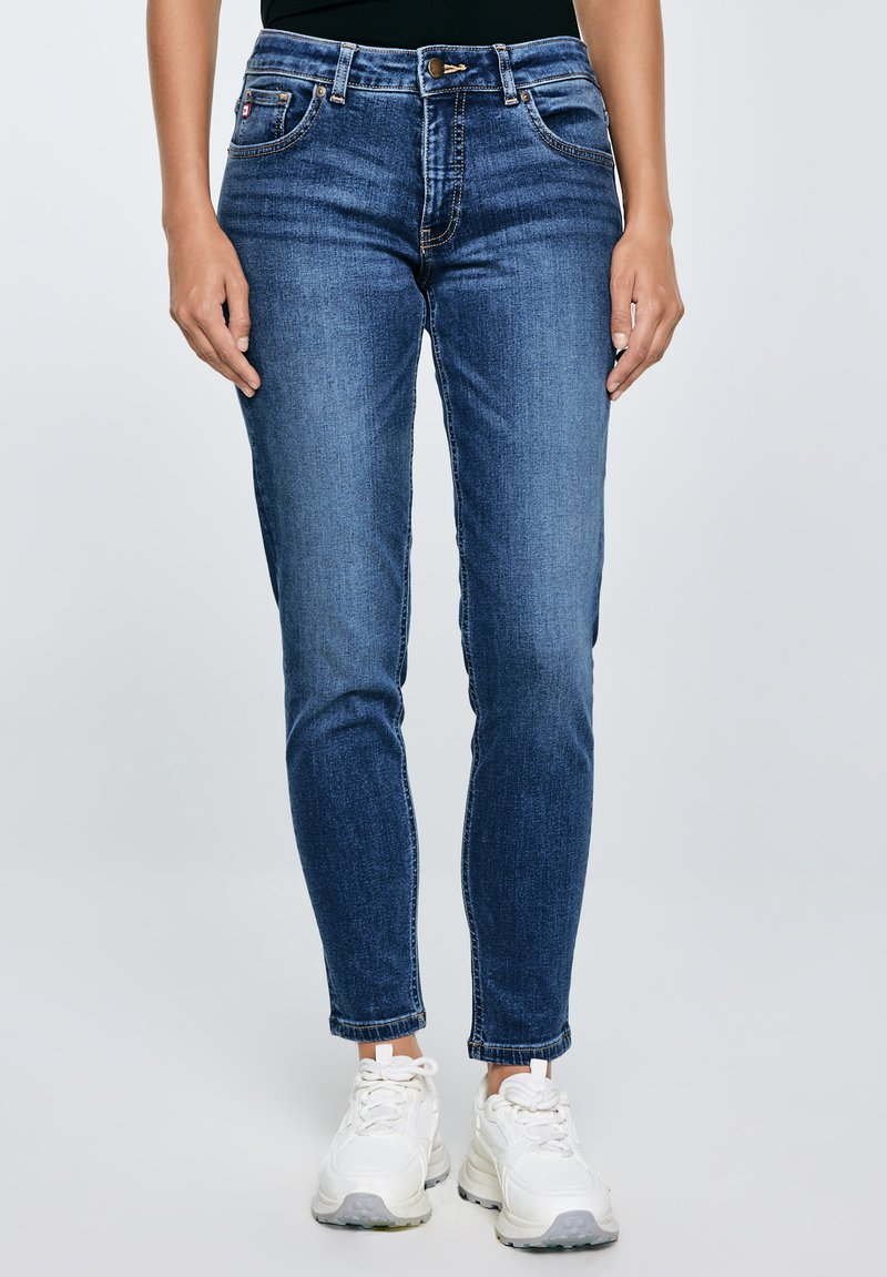 Nő, kék karcsúsított farmernadrágot és fehér tornacipőt visel, egyszerű világos háttér előtt áll.