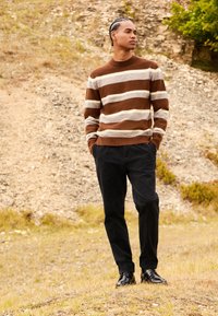 Maglione a righe marroni e crema con scollo rotondo, abbinato a pantaloni scuri e scarpe nere, su uno sfondo esterno con texture.