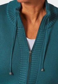 Türkiser gestrickter Pullover mit hohem Kragen, Frontreißverschluss und Kordelzug-Details. Strukturierte Rippstruktur im gesamten Design.