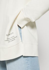 Gros plan d'un sweat-shirt blanc avec une fente latérale et un texte noir indiquant "INGRÉDIENTS : coton biologique, simplicité scandinave, confort facile" sur un jean bleu clair.