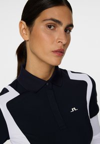 Marinblå polo t-shirt med vita detaljer och en texturerad krage. Har knappslå och en liten logotyp på bröstet. Slät tygfinish.