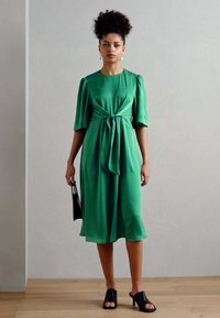Grün, knielanges Kleid aus glattem Stoff, mit einem Gürtel in der Taille und kurzen Puffärmeln; getragen mit schwarzen offenen Mules.